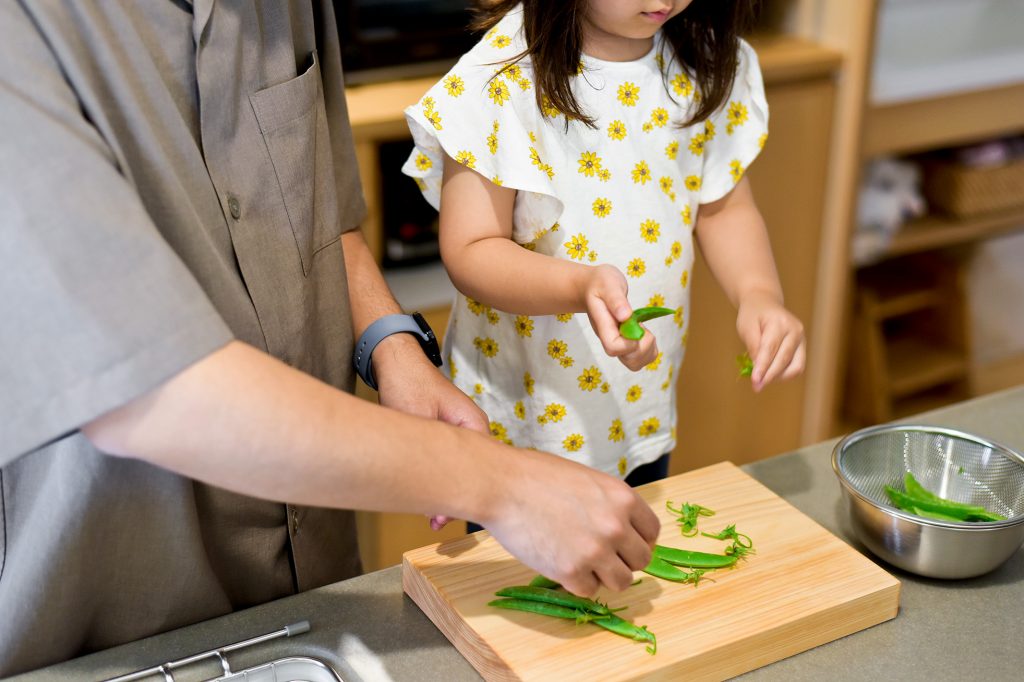 あたたかな住まいのキッチンで、父親と子供が一緒に豆のさやむきをする家族の風景。