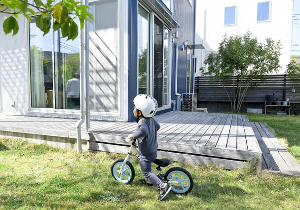 明るい住まいの庭とウッドデッキ。ヘルメットを被った子どもが自転車に乗っている様子。