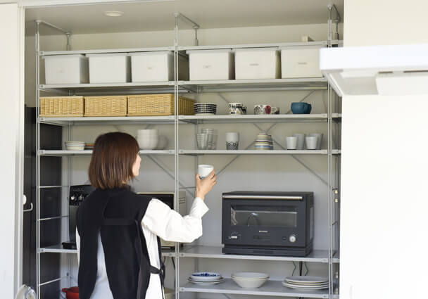 パントリー収納のオープンシェルフと女性。食器や調理家電が整理された明るい収納。