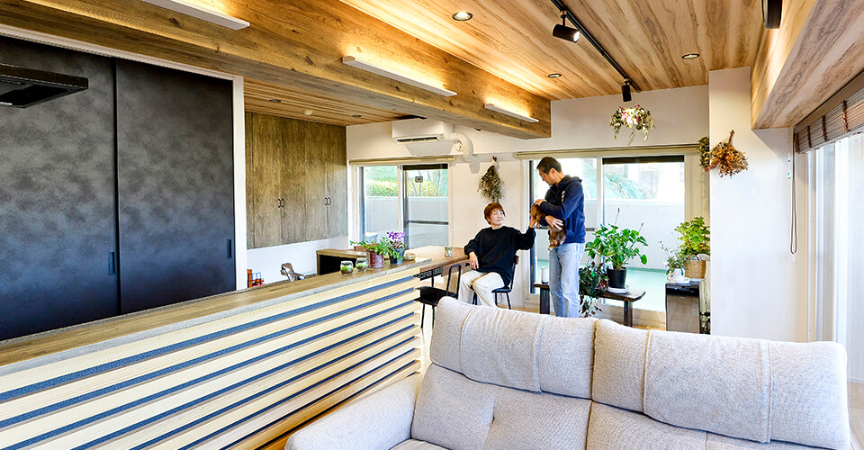 山小屋カフェ風の木製天井とカウンターがあるリビング・キッチンでくつろぐ夫婦と犬
